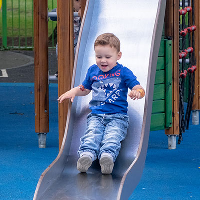 A young toddler boy is sliding down a small steel slide, he is smiling and looking towards the ground.